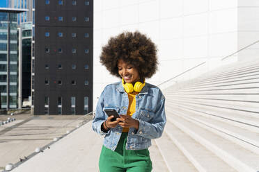 Lächelnde Afro-Frau, die mit ihrem Smartphone Textnachrichten verschickt, steht an einem sonnigen Tag vor einer Treppe - OIPF01796