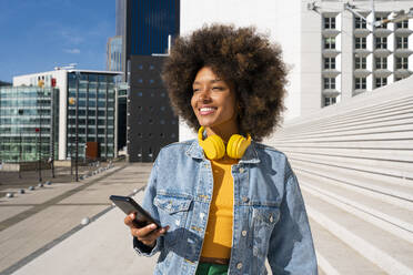 Lächelnde, hübsche junge Frau mit drahtlosen Kopfhörern, die mit einem Mobiltelefon an einer Treppe steht - OIPF01797