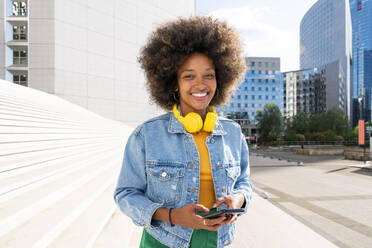 Glückliche junge Frau mit kabellosen Kopfhörern, die mit ihrem Smartphone an einem sonnigen Tag vor einer Treppe steht - OIPF01801