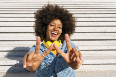 Cheerful Afro woman gesturing peace sign sitting on steps - OIPF01804