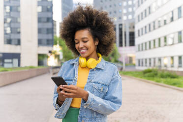 Lächelnde Afro-Frau, die auf dem Fußweg stehend eine Textnachricht über ihr Smartphone sendet - OIPF01811