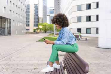 Smiling Afro woman text messaging through smart phone sitting on bench - OIPF01820