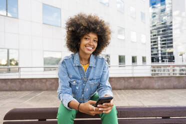 Lächelnde schöne Afro-Frau mit Smartphone sitzt auf Bank - OIPF01822