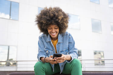 Fröhliche Afro-Frau, die auf einer Bank sitzend Textnachrichten über ihr Smartphone verschickt - OIPF01825