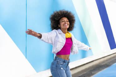 Lächelnde Afro-Frau mit ausgestreckten Armen vor einer blauen Wand - OIPF01859