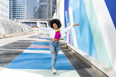 Smiling young woman with arms outstretched walking by colorful wall - OIPF01860