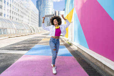 Happy young Afro woman with hands raised walking by colorful wall - OIPF01861