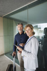 Businesswoman holding mobile phone standing with businessman in front of glass wall - JOSEF10130