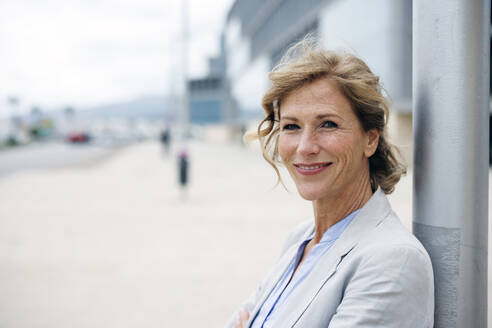 Smiling mature businesswoman standing in front of pole - JOSEF10144