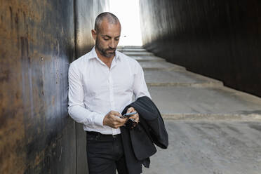 Man using smart phone leaning against wall in alley - JRVF02928
