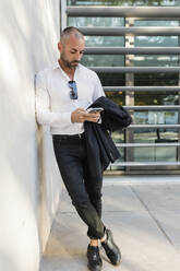 Mann mit Jacke und Smartphone an die Wand gelehnt - JRVF02932