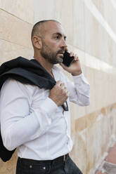 Seriöser Mann mit Jacke, der in der Nähe der Wand mit einem Smartphone spricht - JRVF02935