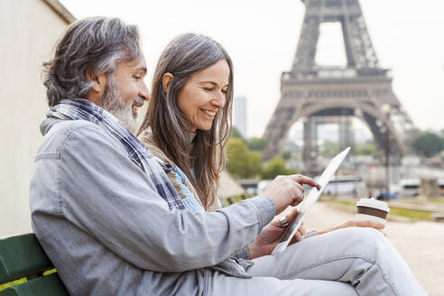 Lächelnde reife Frau mit Mann, der einen Tablet-PC benutzt, auf einer Bank in der Nähe des Eiffelturms, Paris, Frankreich - OIPF01930