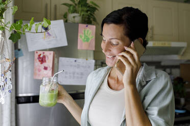 Lächelnde Frau, die ein Saftglas hält und zu Hause mit ihrem Handy telefoniert - TYF00191