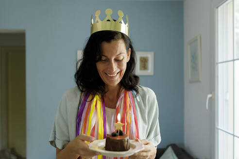 Glückliche Frau mit Krone hält Geburtstagskuchen auf einem Teller zu Hause - TYF00207