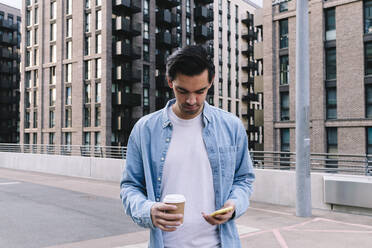 Man holding smart phone and disposable cup standing in front of building - ASGF02342