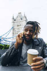 Junger Mann mit Einwegbecher, der mit seinem Smartphone telefoniert, London, England - ASGF02352