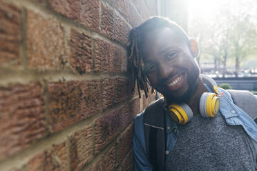 Lächelnder Mann mit drahtlosen Kopfhörern an der Wand lehnend - ASGF02389