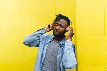 Young man listening music through headphones by yellow wall - ASGF02398