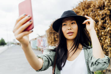 Junge Frau mit Hut macht Selfie mit ihrem Smartphone und strahlt dabei glücklich in die Kamera. - MEUF05893