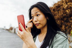 Eine junge Frau mit schwarzen Haaren untersucht ihr Auge, während sie auf ihr Handy schaut - MEUF05911