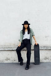 Eine junge Frau sitzt mit ihrem Skateboard an der Wand - MEUF05916