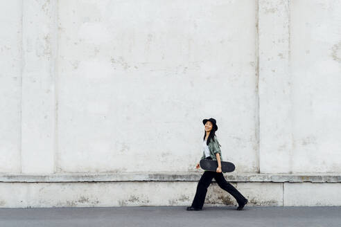 Smiling woman with skateboard walking by wall - MEUF05920