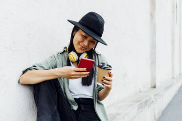 Eine fröhliche Frau sitzt an der Wand und surft mit ihrem Handy im Internet, während sie einen wiederverwendbaren Kaffeebecher hält - MEUF05924