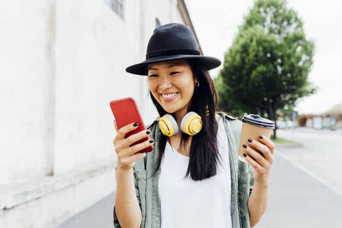 Zufriedene Frau genießt Musik mit kabellosen Kopfhörern, während sie umweltbewusst einen wiederverwendbaren Becher hält und ihr Handy nutzt - MEUF05927