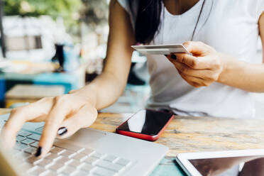 Eine Frau nutzt ihre Kreditkarte, um online in einem Straßencafé zu bezahlen, während ihre Hände auf dem Laptop ruhen - MEUF05935