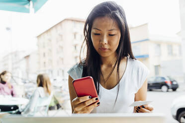 Eine junge Frau bezahlt mit ihrer Kreditkarte online über ihr Smartphone während sie in einem Straßencafé sitzt - MEUF05937