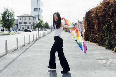 Junge Frau verbreitet Freude mit Regenbogenfahne auf der Straße - MEUF05947