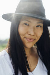 Junge Frau mit strahlendem Lächeln, schwarzem Haar und Hut genießt einen sonnigen Tag - MEUF05951