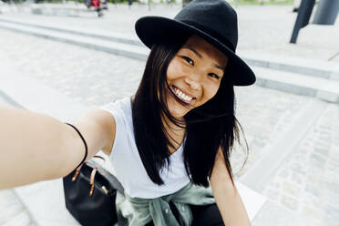 Happy woman with black hair wearing hat taking selfie - MEUF05961