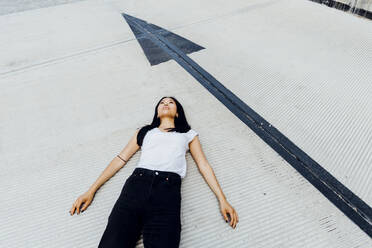 Thoughtful young woman lying by arrow symbol on driveway - MEUF05967