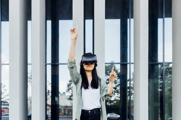 Young woman wearing virtual reality simulator standing with hand raised in front of column - MEUF05977