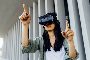 Young woman wearing virtual reality simulator pointing in front of column - MEUF05983