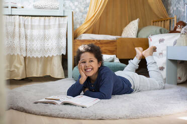 Smiling boy with book looking away while lying down on rug at home - MASF30373