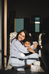 Girl embracing brother while sitting cross-legged in sunlight at home - MASF30399