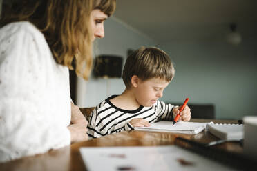 Boy with down syndrome writing in book while sitting by mother at table - MASF30403