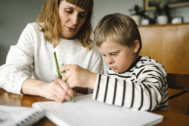 Boy with disability taking support of mother while drawing in book at home - MASF30405