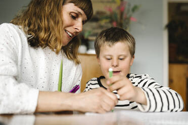 Smiling mother teaching son with disability to draw in book at home - MASF30407