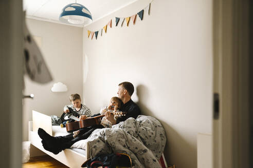 Vater mit Tochter, der seinem behinderten Sohn zu Hause im Schlafzimmer Gitarre beibringt - MASF30411