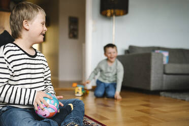 Glücklicher Junge mit Down-Syndrom mit Ball auf dem Boden sitzend im Wohnzimmer - MASF30436