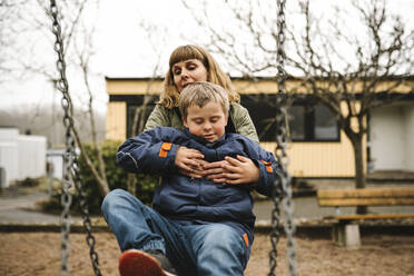 Son with down syndrome taking help of mother while sitting in swing at park - MASF30446