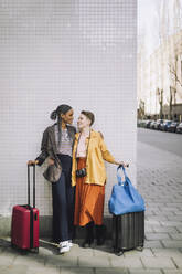Full length of smiling lesbian couple with wheeled luggage standing against wall - MASF30472
