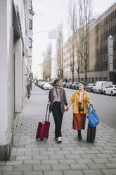 Lesbian couple holding hands while walking with wheeled luggage on sidewalk - MASF30473