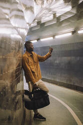 Junger Mann in voller Länge, der eine Tasche trägt, während er ein Selfie mit seinem Mobiltelefon in einem U-Bahn-Tunnel macht - MASF30510