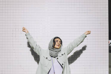 Smiling woman with arms raised standing against wall during sunny day - MASF30571