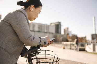 Seitenansicht einer Geschäftsfrau mit In-Ear-Kopfhörern, die ein Smartphone benutzt, während sie sich an ein Fahrrad lehnt - MASF30602